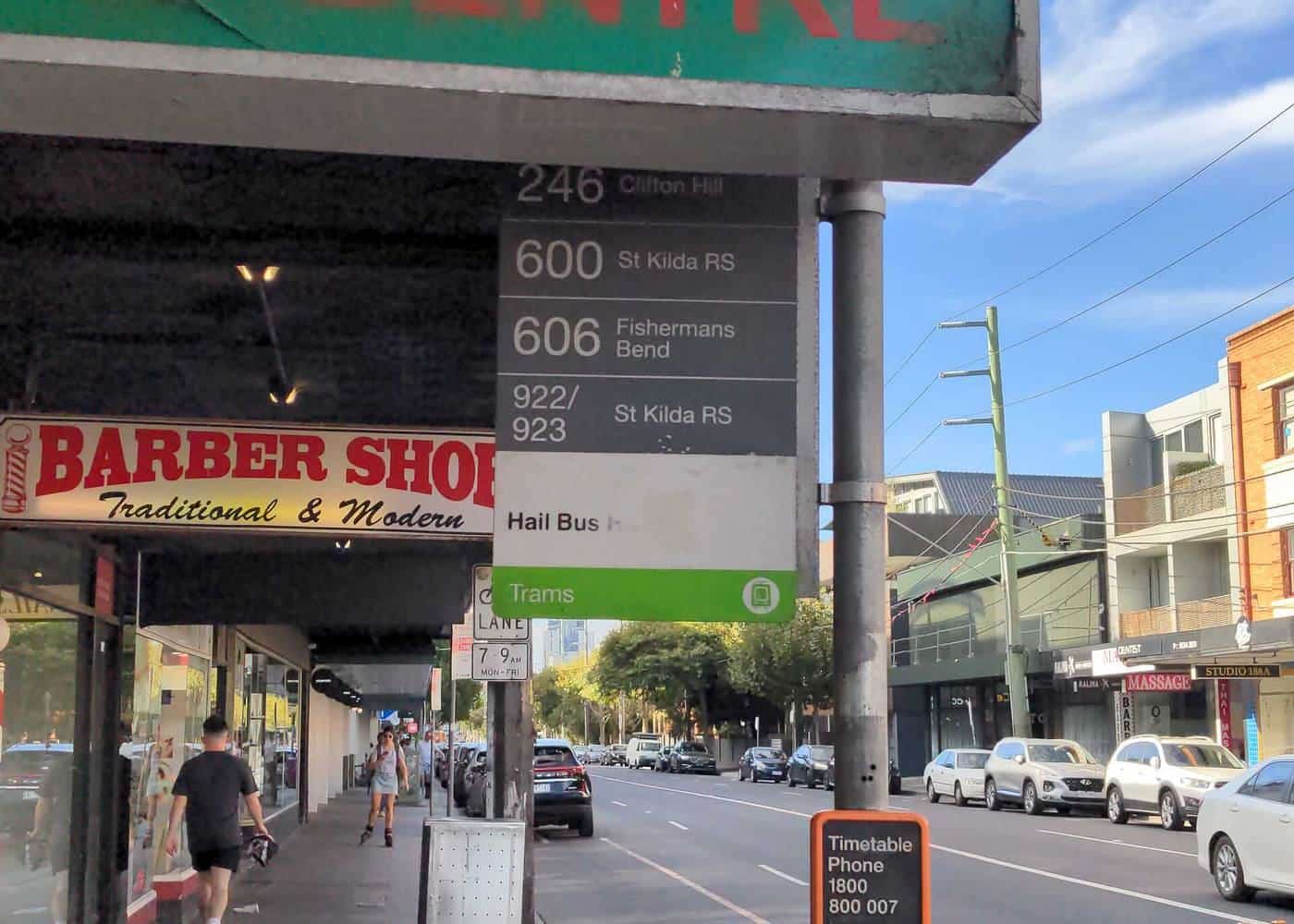 A shop sign mounted on a pole on a busy St Kilda street partly blocking a bus stop sign behind it